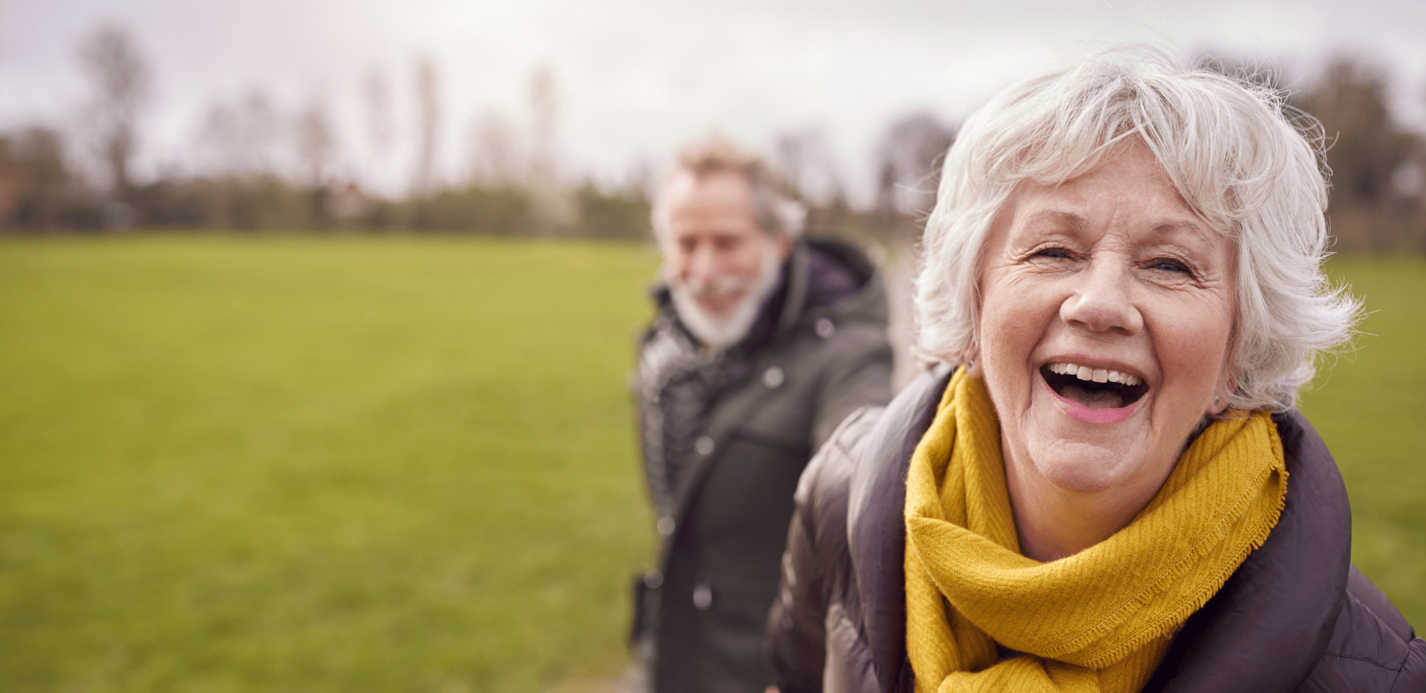 A joyful elderly woman smiling in the foreground with an older man out of focus in the background outdoors.