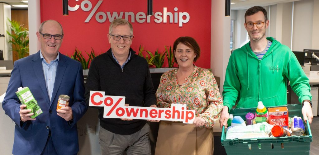 Four colleagues smiling in an office environment, holding a sign and groceries to represent co-ownership.