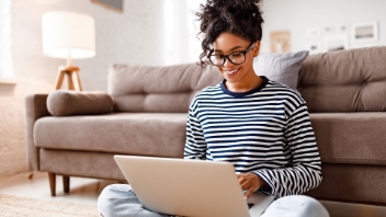 A woman using a laptop while sitting on the floor leaning against a sofa.