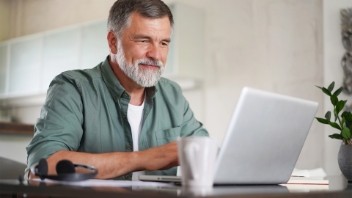 A mature man with a beard smiling while working on a laptop at a home office desk.